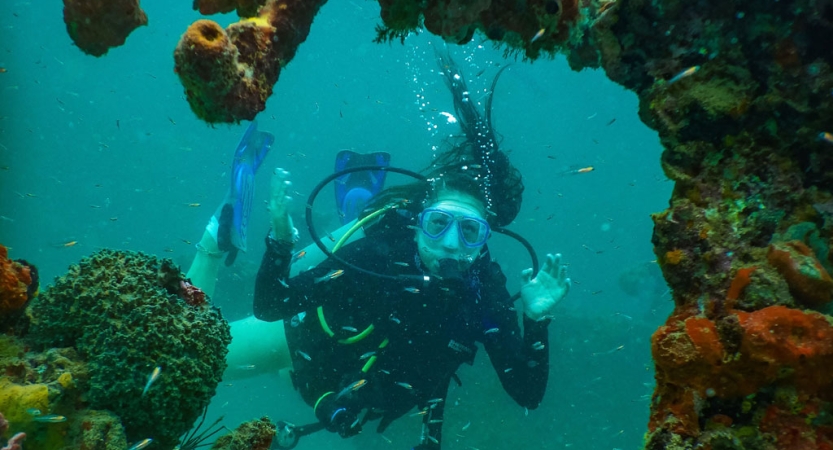 A person who is scuba diving underwater looks at the camera through a hole in coral reef. 